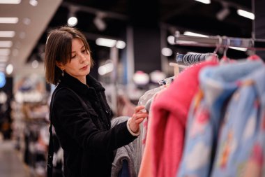 Siyah ceketli şık bir kadın giyim mağazasında yumuşak, renkli kazaklara göz gezdiriyor ve gardırobu için dikkatlice seçiyor..