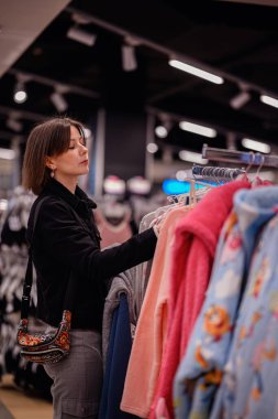 Siyah ceketli şık bir kadın giyim mağazasında yumuşak, renkli kazaklara göz gezdiriyor ve gardırobu için dikkatlice seçiyor..