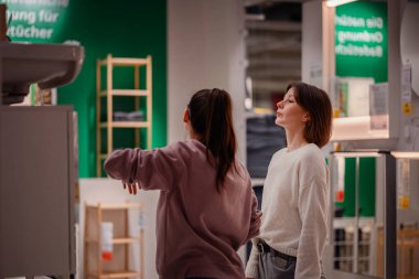 İki kadın bir mağazaya girip banyo bölümündeki lavabo dolabını inceliyor. Biri ayrıntıları incelemek için uzanırken diğeri gözlemliyor..