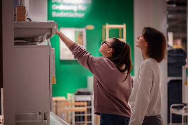 İki kadın bir mağazaya girip banyo bölümündeki lavabo dolabını inceliyor. Biri ayrıntıları incelemek için uzanırken diğeri gözlemliyor..