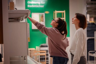 İki kadın bir mağazaya girip banyo bölümündeki lavabo dolabını inceliyor. Biri ayrıntıları incelemek için uzanırken diğeri gözlemliyor..