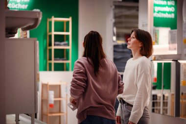 İki kadın bir mağazaya girip banyo bölümündeki lavabo dolabını inceliyor. Biri ayrıntıları incelemek için uzanırken diğeri gözlemliyor..