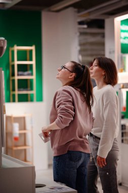 İki kadın bir mağazaya girip banyo bölümündeki lavabo dolabını inceliyor. Biri ayrıntıları incelemek için uzanırken diğeri gözlemliyor..