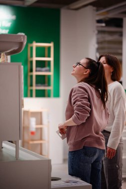 İki kadın bir mağazaya girip banyo bölümündeki lavabo dolabını inceliyor. Biri ayrıntıları incelemek için uzanırken diğeri gözlemliyor..