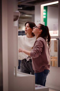 İki kadın ev dekorasyonu mağazasında bir banyo aksesuarını incelerken, ev dekorasyonu malzemelerini ararken ürün detaylarını tartışıyorlar..