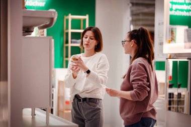 İki kadın ev dekorasyonu mağazasında bir banyo aksesuarını incelerken, ev dekorasyonu malzemelerini ararken ürün detaylarını tartışıyorlar..