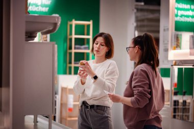 İki kadın ev dekorasyonu mağazasında bir banyo aksesuarını incelerken, ev dekorasyonu malzemelerini ararken ürün detaylarını tartışıyorlar..