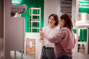 İki kadın ev dekorasyonu mağazasında bir banyo aksesuarını incelerken, ev dekorasyonu malzemelerini ararken ürün detaylarını tartışıyorlar..