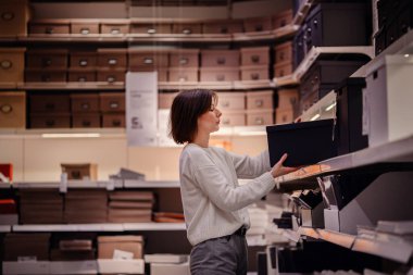 Beyaz kazaklı bir kadın, depolama seçeneklerini düzenlerken bir de kara kutu seçen bir ev dükkanındaki kutuları karıştırıyor..