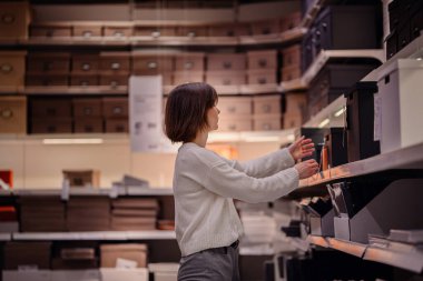 Beyaz kazaklı bir kadın, depolama seçeneklerini düzenlerken bir de kara kutu seçen bir ev dükkanındaki kutuları karıştırıyor..