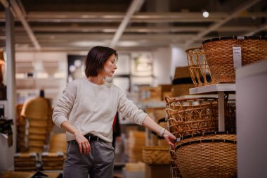 Beyaz kazaklı bir kadın bir mağazada dokuma sepetleri karıştırıyor, depo veya dekor için dikkatlice bir tane seçiyor. Sepetlerin dokusunu ve boyutunu inceliyor..
