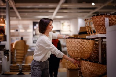 Beyaz kazaklı bir kadın bir mağazada dokuma sepetleri karıştırıyor, depo veya dekor için dikkatlice bir tane seçiyor. Sepetlerin dokusunu ve boyutunu inceliyor..