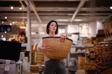 Beyaz kazaklı bir kadın, ev eşyaları alırken depolama veya dekor için bir mağazada büyük bir dokuma sepetini inceliyor..