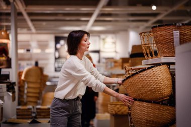 Beyaz kazaklı bir kadın bir mağazada dokuma sepetleri karıştırıyor, depo veya dekor için dikkatlice bir tane seçiyor. Sepetlerin dokusunu ve boyutunu inceliyor..