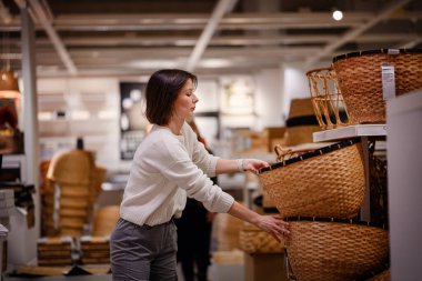 Beyaz kazaklı bir kadın bir mağazada dokuma sepetleri karıştırıyor, depo veya dekor için dikkatlice bir tane seçiyor. Sepetlerin dokusunu ve boyutunu inceliyor..