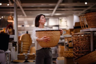 Beyaz kazaklı bir kadın, ev eşyaları alırken depolama veya dekor için bir mağazada büyük bir dokuma sepetini inceliyor..