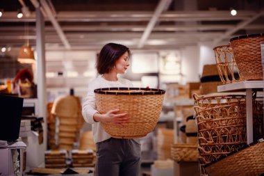 Beyaz kazaklı bir kadın, ev eşyaları alırken depolama veya dekor için bir mağazada büyük bir dokuma sepetini inceliyor..