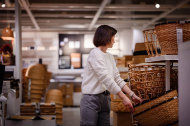 Beyaz kazaklı bir kadın bir mağazada dokuma sepetleri karıştırıyor, depo veya dekor için dikkatlice bir tane seçiyor. Sepetlerin dokusunu ve boyutunu inceliyor..
