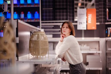 Beyaz kazaklı bir kadın, dekoratif bir lambayı düşünceli bir şekilde rafa yaslanıyor ve bir mağazada ışıklandırma için alışveriş yapıyor..