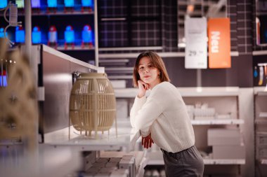 Beyaz kazaklı bir kadın, dekoratif bir lambayı düşünceli bir şekilde rafa yaslanıyor ve bir mağazada ışıklandırma için alışveriş yapıyor..