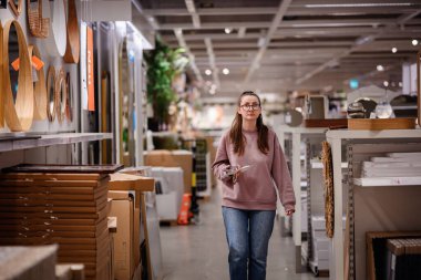 Pembe kazaklı bir kadın bir mobilya mağazasında ev dekorasyonu ve depolama raflarına göz atar ve bir şey seçerken telefonunu tutar..