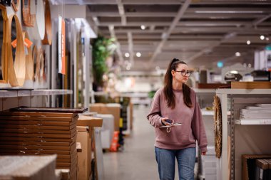 Pembe kazaklı bir kadın bir mobilya mağazasında ev dekorasyonu ve depolama raflarına göz atar ve bir şey seçerken telefonunu tutar..