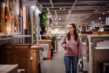 Pembe kazaklı bir kadın bir mobilya mağazasında ev dekorasyonu ve depolama raflarına göz atar ve bir şey seçerken telefonunu tutar..