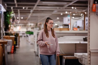 Pembe kazaklı bir kadın bir mobilya mağazasında ev dekorasyonu ve depolama raflarına göz atar ve bir şey seçerken telefonunu tutar..