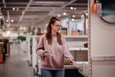 Pembe kazaklı bir kadın bir mobilya mağazasında ev dekorasyonu ve depolama raflarına göz atar ve bir şey seçerken telefonunu tutar..