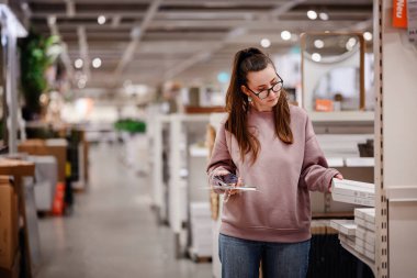 Pembe kazaklı bir kadın bir mobilya mağazasında ev dekorasyonu ve depolama raflarına göz atar ve bir şey seçerken telefonunu tutar..