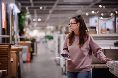 Pembe kazaklı bir kadın bir mobilya mağazasında ev dekorasyonu ve depolama raflarına göz atar ve bir şey seçerken telefonunu tutar..