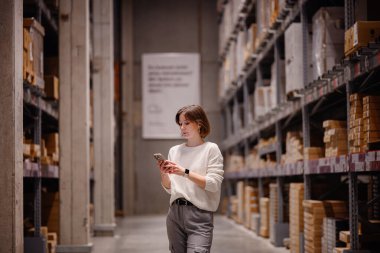 Beyaz kazaklı bir kadın depo koridorunda yürürken, akıllı telefonunu kontrol ederken, paketlenmiş rafları ya da mobilyaları karıştırıyor..