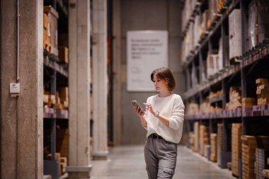 Beyaz kazaklı bir kadın depo koridorunda yürürken, akıllı telefonunu kontrol ederken, paketlenmiş rafları ya da mobilyaları karıştırıyor..