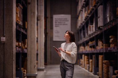 Beyaz kazaklı bir kadın depo koridorunda yürürken, akıllı telefonunu kontrol ederken, paketlenmiş rafları ya da mobilyaları karıştırıyor..