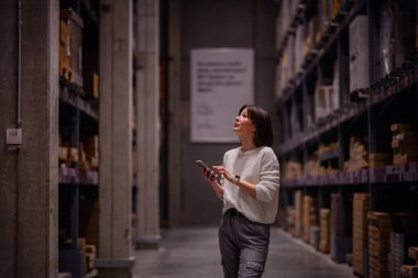 Beyaz kazaklı bir kadın depo koridorunda yürürken, akıllı telefonunu kontrol ederken, paketlenmiş rafları ya da mobilyaları karıştırıyor..