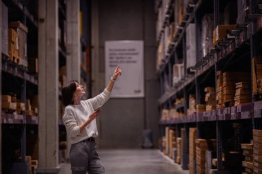 Beyaz kazaklı bir kadın depo koridorunda geziniyor, akıllı telefonunu kontrol ederken yüksek bir rafı işaret ediyor, muhtemelen bir ürün arıyor..