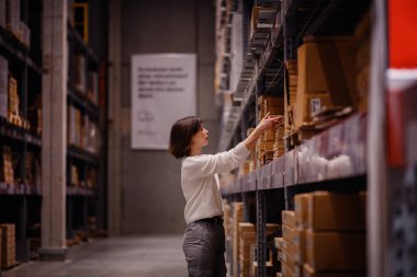 Beyaz kazaklı bir kadın bir depo koridorunda geziniyor, yüksek bir raftaki bir ürüne uzanırken ev eşyası veya mobilya alışverişi yapıyor..