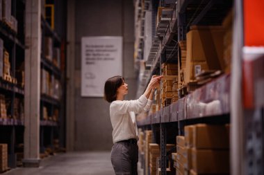 Beyaz kazaklı bir kadın bir depo koridorunda geziniyor, yüksek bir raftaki bir ürüne uzanırken ev eşyası veya mobilya alışverişi yapıyor..