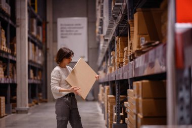 Beyaz kazaklı bir kadın depo tarzı bir mobilya mağazasında alışveriş yaparken büyük bir düz paket kutusu alıyor ve ev dekorasyonu için bir eşya seçiyor..
