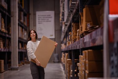Beyaz kazaklı bir kadın depo tarzı bir mobilya mağazasında alışveriş yaparken büyük bir düz paket kutusu alıyor ve ev dekorasyonu için bir eşya seçiyor..