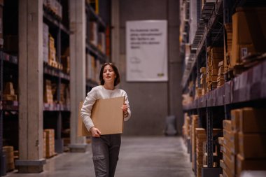 Beyaz kazaklı bir kadın depo tarzı bir mobilya mağazasında alışveriş yaparken büyük bir düz paket kutusu alıyor ve ev dekorasyonu için bir eşya seçiyor..