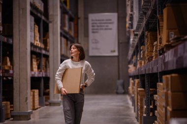 Beyaz kazaklı bir kadın depo tarzı bir mobilya mağazasında alışveriş yaparken büyük bir düz paket kutusu alıyor ve ev dekorasyonu için bir eşya seçiyor..