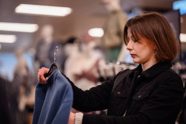 Bir kadın bir giyim mağazasında alışveriş yaparken mavi bir kazağı dikkatle inceler. Odaklanmış görünüyor, mağazanın sıcak ışıklandırmasının altındaki kumaşı ve stili kontrol ediyor..