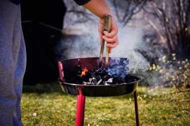 Güneşli bir arka bahçede ızgarada sigara içmek için ahşap maşa kullanan biri..