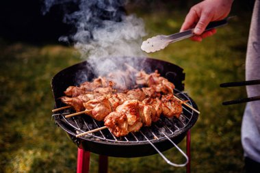 Kömür ızgarasında pişen sulu salamura edilmiş et şişleri. Birisi onlara maşa kullanırken duman yükseliyor..