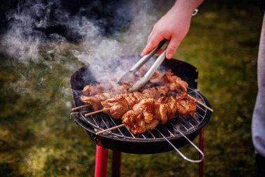 Kömür ızgarasında pişen sulu salamura edilmiş et şişleri. Birisi onlara maşa kullanırken duman yükseliyor..
