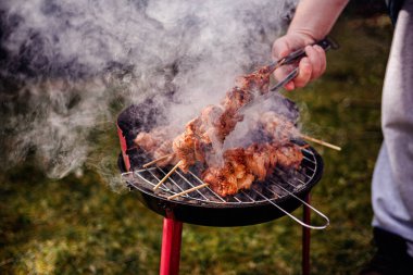 Kömür ızgarasında pişen sulu salamura edilmiş et şişleri. Birisi onlara maşa kullanırken duman yükseliyor..
