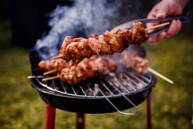 Arka planda dumanlar yükselirken kömür mangalından ızgara et şişini kaldıran bir elin yakın çekimi..