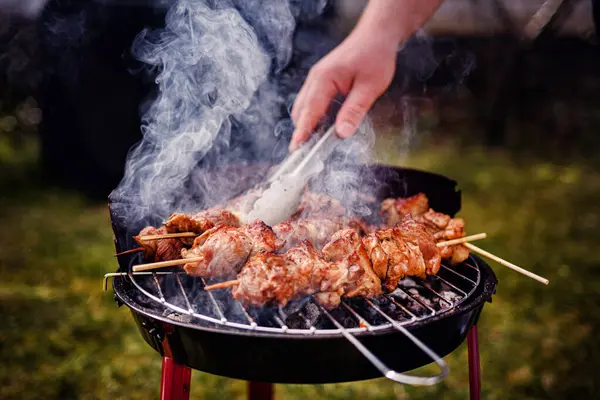 Kömür ızgarasında pişen sulu salamura edilmiş et şişleri. Birisi onlara maşa kullanırken duman yükseliyor..