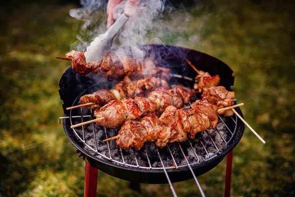 Kömür ızgarasında pişen sulu salamura edilmiş et şişleri. Birisi onlara maşa kullanırken duman yükseliyor..